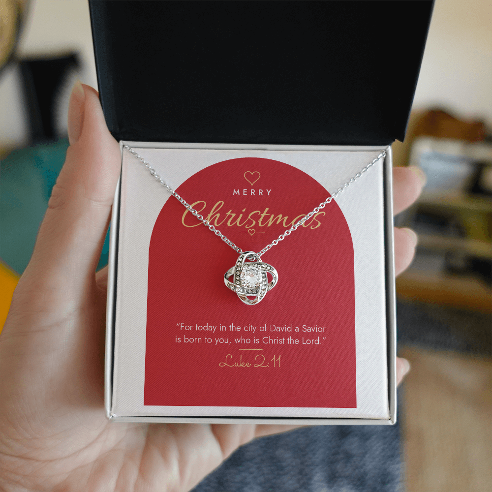 Hand holding open gift box with 14K white gold finish Christian Christmas Love Knot necklace and Luke 2:11 modern message card