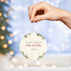 Hand hanging acrylic bulb ornament with Our First Christmas Mr. & Mrs. 2026 design in front of Christmas tree lights